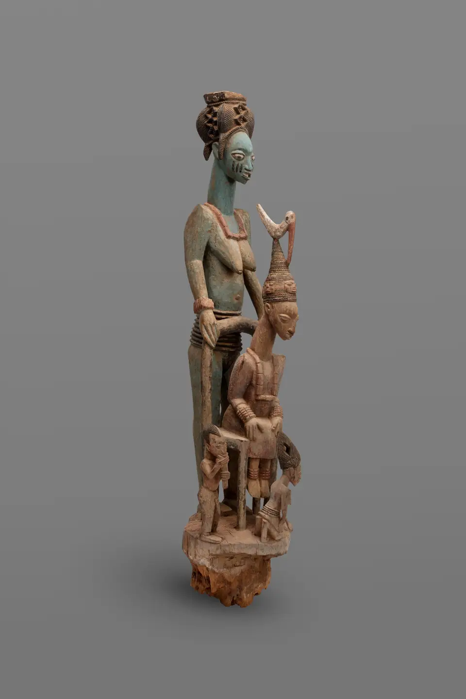 A tall carved wooden post of a seated king with a woman standing behind him and smaller figures around.