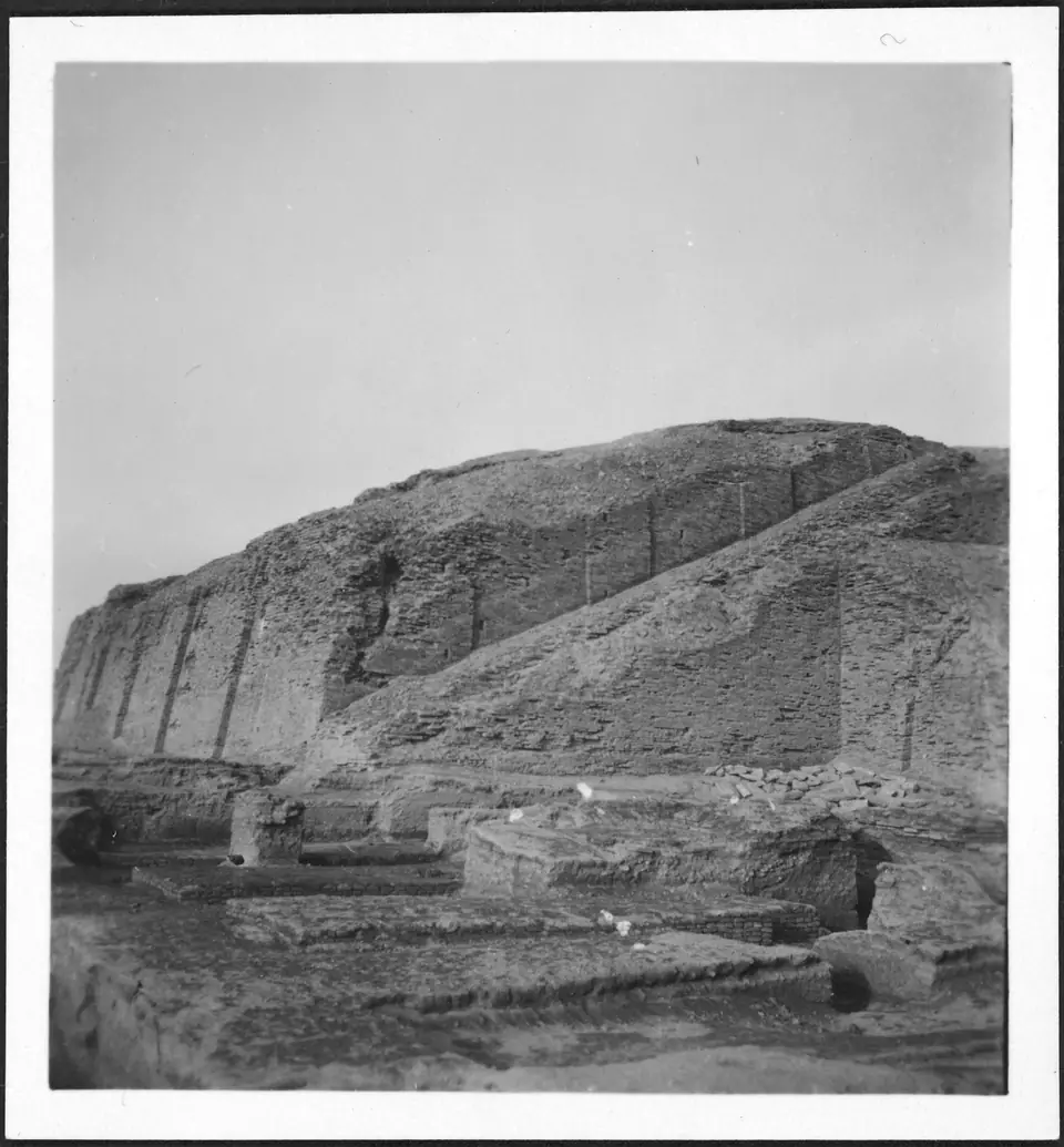 An old black-and-white photograph of a massive mud-brick platform with sloping walls and a long ramp up its side, above excavated foundations.