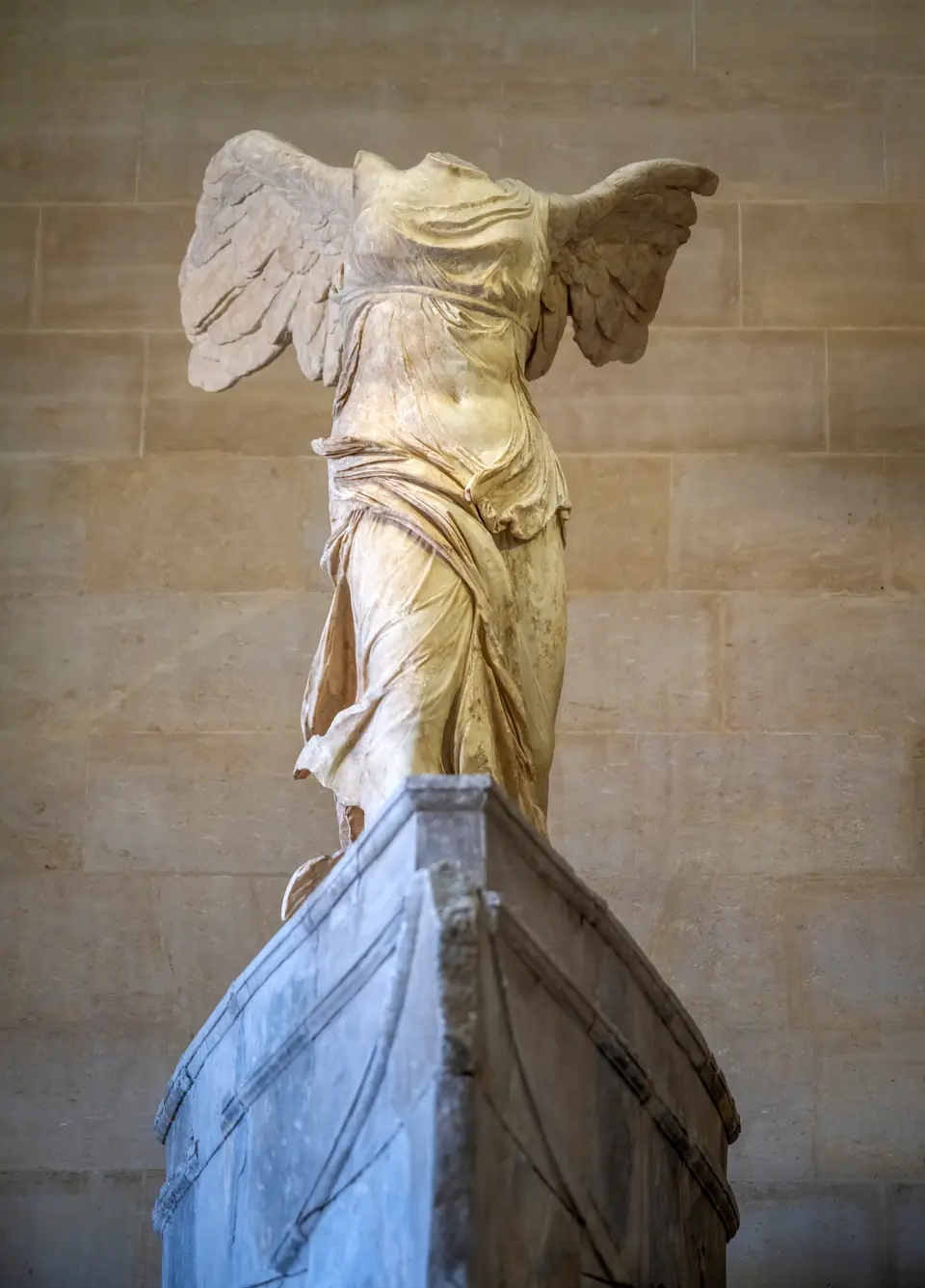 A headless marble statue of a winged woman striding forward, her thin drapery pressed against her body by the wind.